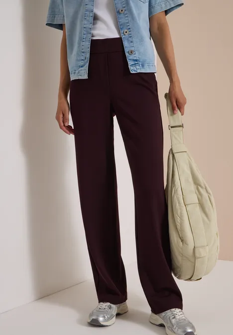Frau trägt plumfarbene weite Hose, helle Jeansjacke, silberne Sneaker und hält einen beigen Stoffbeutel.
