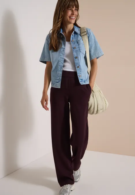 Frau in burgunderroter Marlene-Hose, weißem Top, jeansblauer Kurzjacke und Sneakers mit Tasche.
