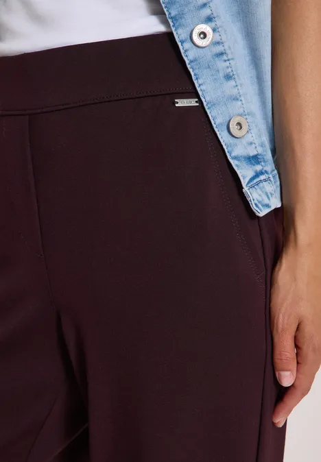 Nahaufnahme einer Person in weinroter Hose, weißem Oberteil und Jeansjacke mit Hand an der Seite