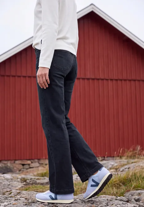 Personne en veste blanche, jean foncé et baskets blanches devant un bâtiment en bois rouge.