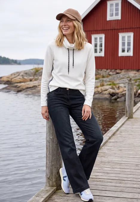 Femme en sweat blanc, jeans noirs évasés et baskets blanches sur un ponton devant une maison rouge au bord de l'eau.