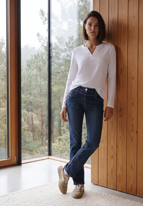 High Waist Bootcut Jeans im Slim Fit Dark blue wash Eine Frau steht lässig in Jeans und weißer Bluse an einer Holzwand neben einem Fenster mit Waldblick.