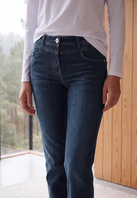 High Waist Bootcut Jeans im Slim Fit Dark blue wash Eine Person in dunkelblauen Jeans und weißem Oberteil steht vor einem Fenster neben einer Holzverkleidung.