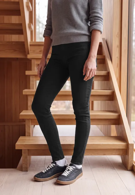 High Waist Skinny Leg Jeggings im Slim Fit Black Eine Person in schwarzer Skinny-Jeans, grauem Pullover und schwarzen Sneakern steht auf einer Holztreppe.