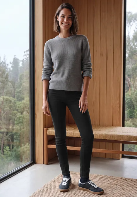 High Waist Skinny Leg Jeggings im Slim Fit Black Frau in grauem Pullover, schwarzer Hose und Sneakers steht lächelnd vor großem Fenster in modernem Holzraum.