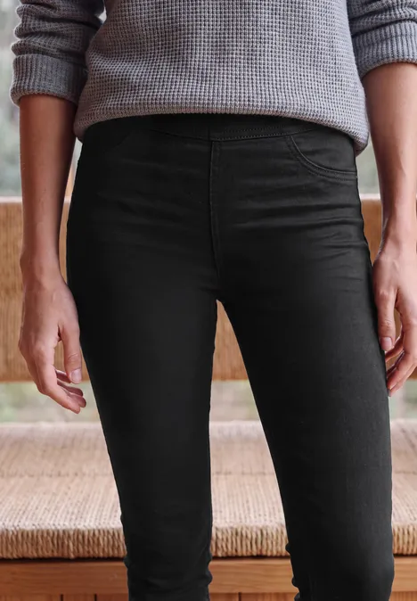 High Waist Skinny Leg Jeggings im Slim Fit Black Eine Person trägt schwarze enge Hose und grauen Pullover, von der Taille bis zu den Knien.