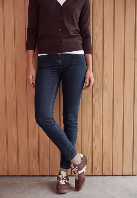 High Waist Skinny Leg Jeggings im Slim Fit rinsed wash Unterkörper einer Person in dunklen Skinny-Jeans, braunen Sneakern und Cardigan vor Holzverkleidung.