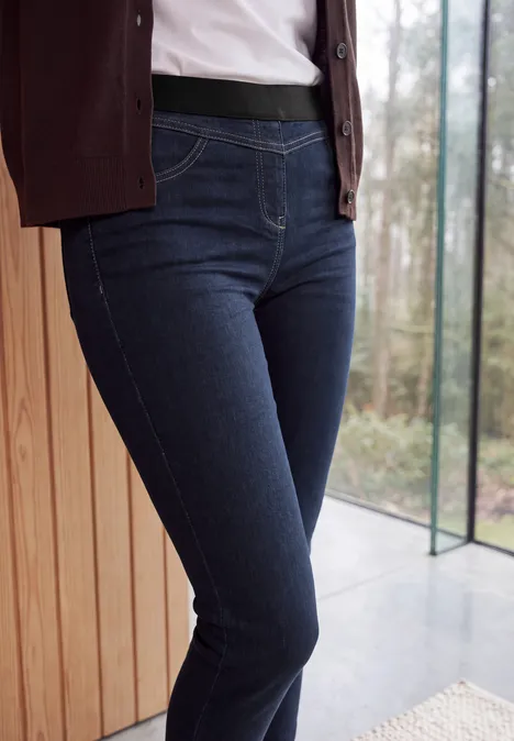 High Waist Skinny Leg Jeggings im Slim Fit rinsed wash Eine Person trägt dunkle Jeans und steht vor einem Fenster neben einer Holzverkleidung.