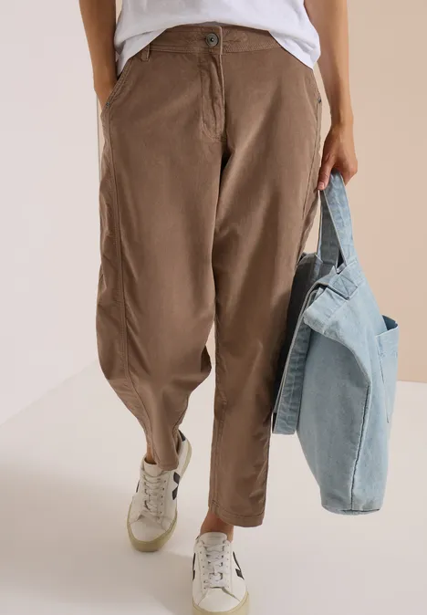 Person trägt lockere braune Hose, weiße Sneakers und hält eine Jeansjacke in der Hand.