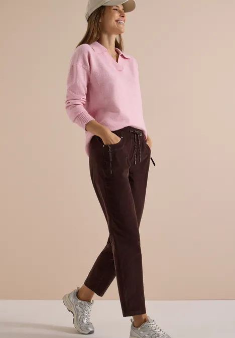 Mid Waist Slim Leg Hose im Casual Fit truffle brown Eine sportliche Frau in rosa Pullover, braunen Hosen und Mütze, die lässig in weißen Sneakern steht.