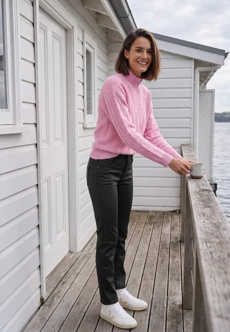 Mid Waist Straight Leg Hose im Slim Fit Black Frau in rosa Pullover, schwarzer Hose und weißen Stiefeletten lehnt auf einer Holzterrasse am Wasser und lächelt.