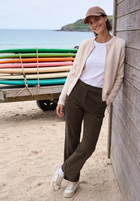 Eine lächelnde Frau lehnt entspannt an einem Holzhaus am Strand, im Hintergrund bunte Surfbretter.