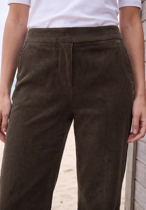 Frau trägt hoch taillierte, dunkelbraune Cordhose mit weißem T-Shirt, Hände locker seitlich.