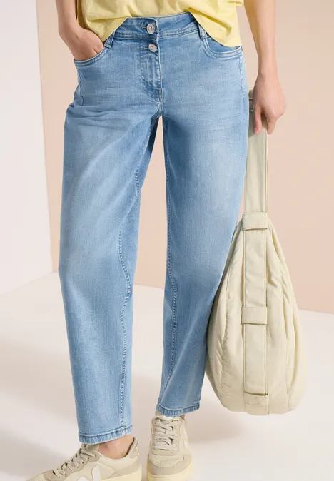 Frau in hellblauen Jeans, gelbem Top, beigen Sneakern trägt eine beige Tasche.