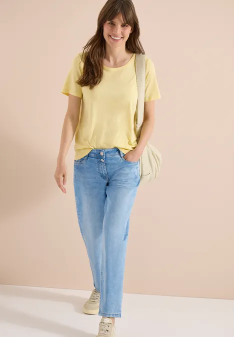 Eine Frau trägt ein gelbes T-Shirt, helle Jeans und Sneakers, mit Schultertasche vor neutralem Hintergrund.