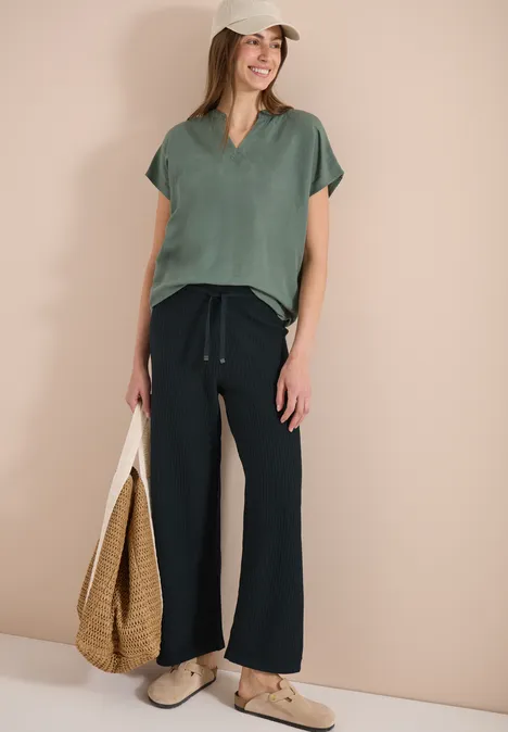Frau in grüner Bluse, schwarzen Hosen, weißen Schuhen mit Kappe und Basttasche vor beiger Wand.
