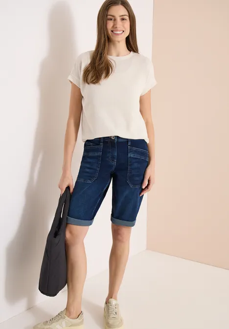 Vrouw met wit T-shirt en donker denim bermudashorts, draagt tas en sneakers.