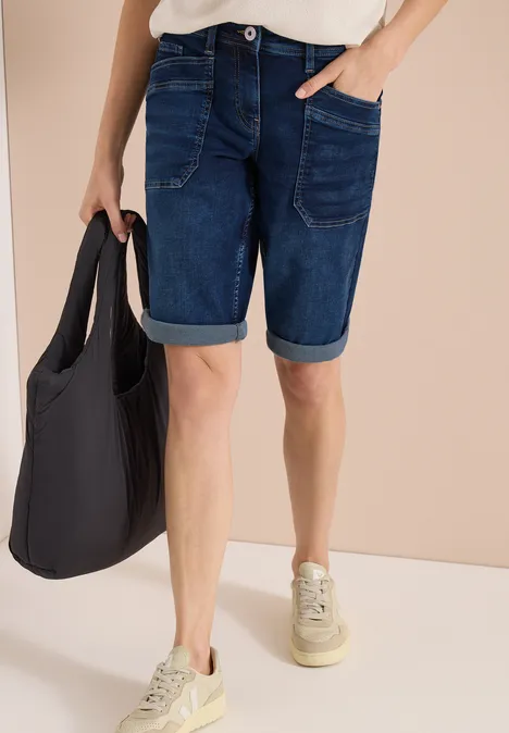 Persoon met donkere jeansshorts, witte sneakers en een zwarte stoffen tas.