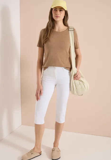 Eine Frau trägt ein beiges T-Shirt, weiße Caprihose, beige Schuhe und hat eine passende Umhängetasche.