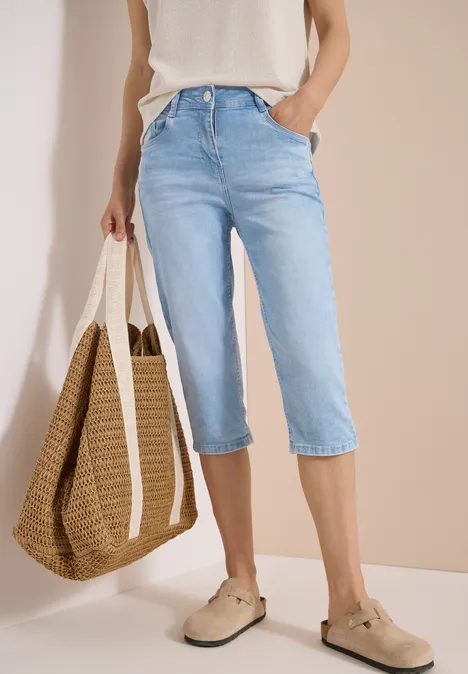 High Waist Slim Leg Jeans im Slim Fit light blue random bleached Frau in hellblauer Capri-Jeans mit weißem Oberteil, Basttasche und beigen Espadrilles vor neutralem Hintergrund.