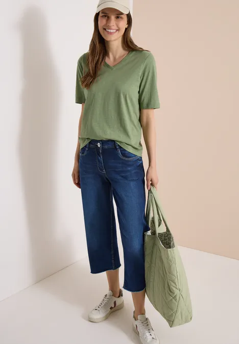 Frau trägt grünes V-Shirt, weite blaue Capri-Jeans, weiße Sneaker und große grüne Tasche vor hellem Hintergrund.