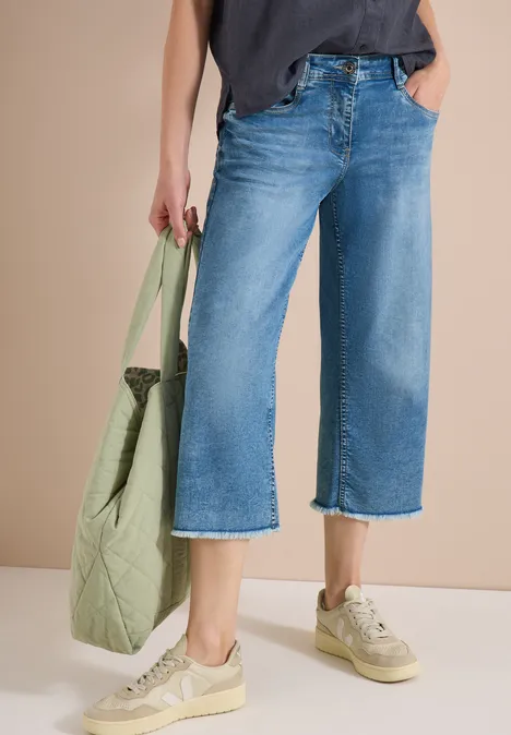 Frau trägt weite, knöchellange Jeans, beige Sneaker und hält eine grüne, gesteppte Tasche.