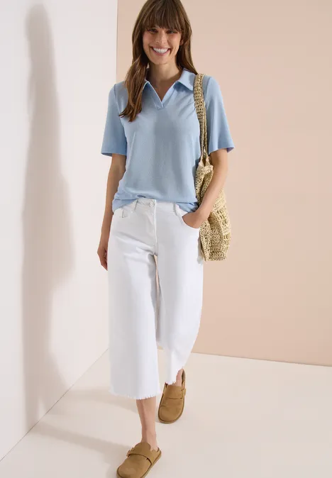 Frau mit hellblauem Poloshirt, weißen Culotte-Hosen, braunen Schuhen und Strohtasche vor schlichter Kulisse.