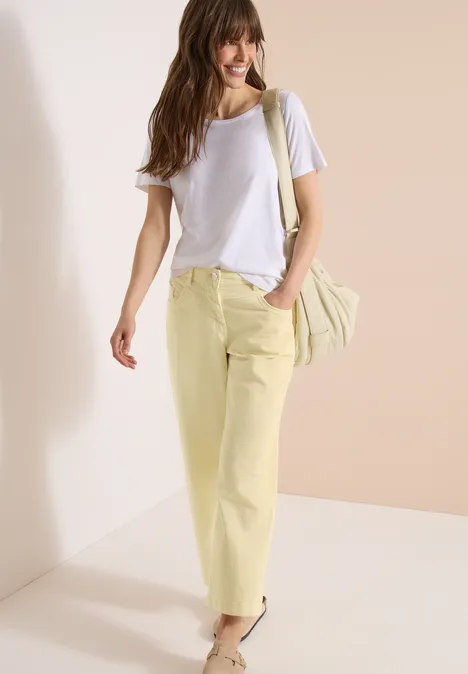 Frau trägt lässiges Outfit mit weißem T-Shirt, gelben Hosen und Umhängetasche vor hellem Hintergrund.