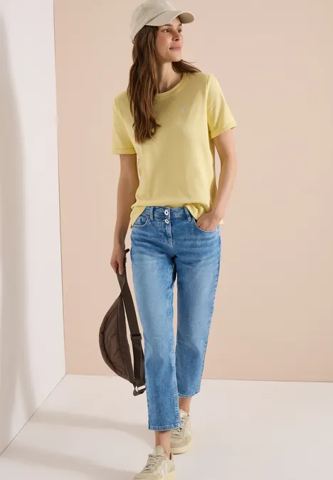 Vrouw in gele T-shirt, lichtblauwe spijkerbroek, sneakers en pet, met een tas los over de schouder.