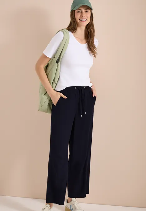 Eine Frau in weißem T-Shirt, weiter dunkelblauer Hose mit Kordelzug und grüner Umhängetasche vor beigem Hintergrund.