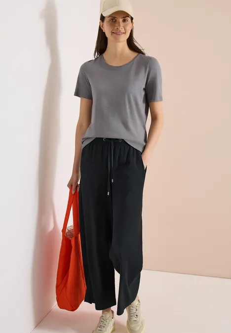 Femme en t-shirt gris, pantalon ample noir et baskets, tenant un sac rouge.