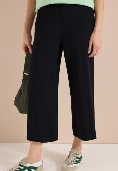 Vrouw draagt zwarte, wijde enkellange broek met witte sneakers en een kaki tas.