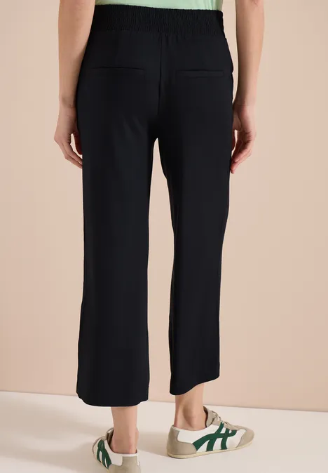 Achteraanzicht van een persoon met zwarte, wijde broek en witte sneakers met groene strepen voor een beige achtergrond.