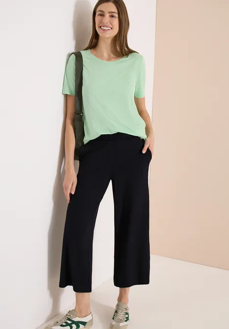 Vrouw tegen lichte muur, draagt mintgroen t-shirt, zwarte wijde culottes en sandalen.