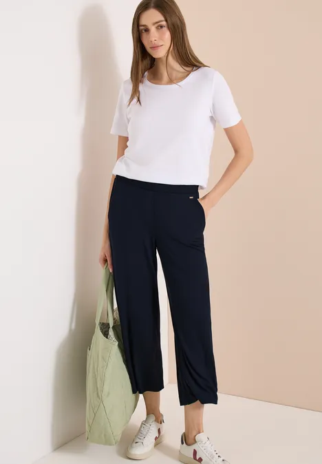 Frau in weißem T-Shirt, navy Capri-Hose, weißen Sneakers, hält grünen Stoffbeutel.