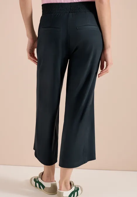 Vrouw van achteren in loszittende zwarte cropped broek en groene sandalen.