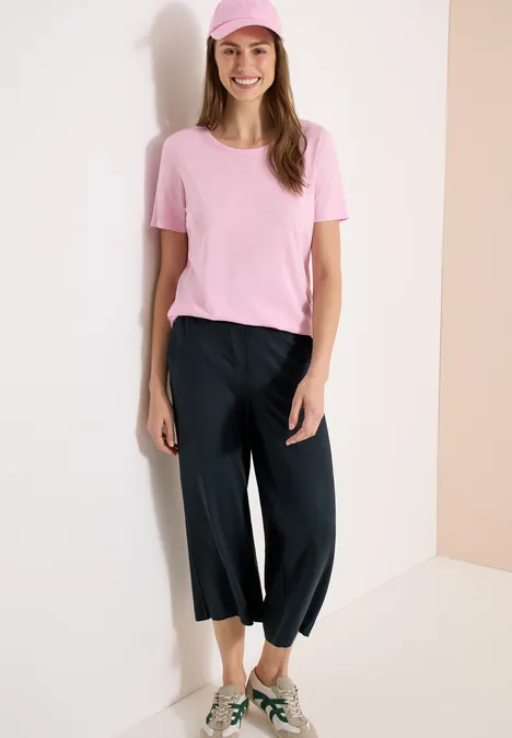 Vrouw in roze T-shirt en losse zwarte driekwartbroek, staand voor een witte muur in een hoek.