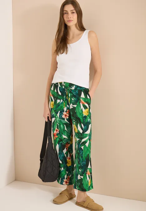 High Waist Wide Leg Hose im Loose Fit khaki Frau trägt weißes Tanktop, grüne Palmenprint-Hose, beigen Hintergrund, schwarze Tasche und beigen Sandalen.