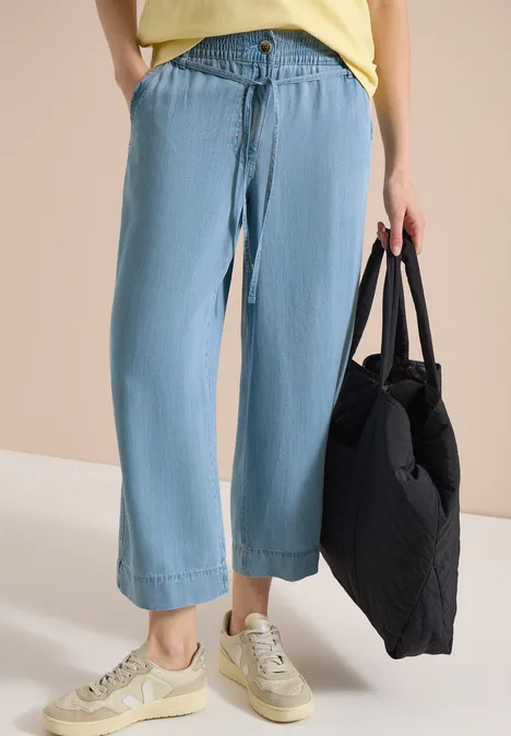 Eine Person trägt weite, hellblaue Jeans, beige Sneaker und hält eine schwarze Tasche.