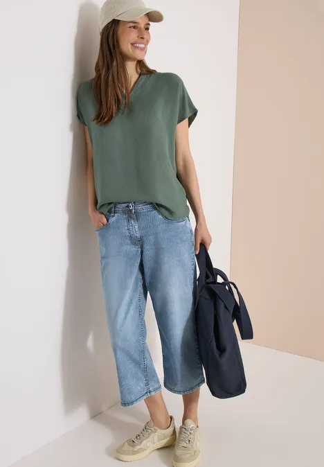 Femme décontractée appuyée contre un mur, t-shirt vert, pantalon ample en jean, baskets et sac.