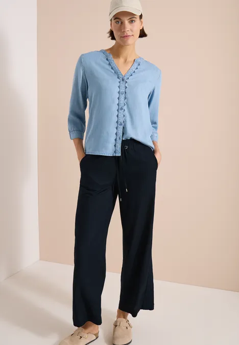 Wide Leg Hose mit tonalem Tape-Detail universal blue Modell trägt hellblaues, vorn geknöpftes Oberteil mit Rüschen und weite schwarze Hosen mit Sneakers.
