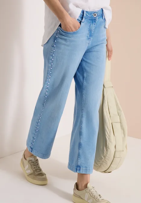 Lichtblauwe, rechte jeans met sierknopen langs de pijpen, gecombineerd met beige sneakers en tas.