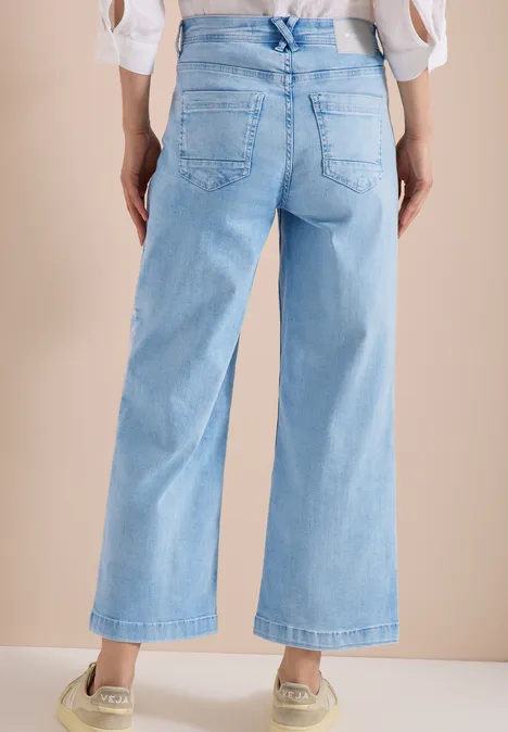 Achteraanzicht van een persoon met lichtblauwe, wijde, enkellange jeans en sneakers tegen een beige achtergrond.