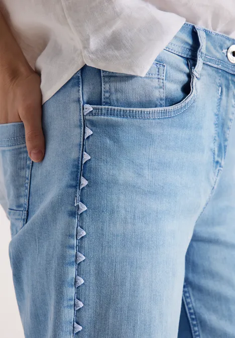 Lichtblauwe jeans met driehoekdetails langs de zijnaad, witte blouse en hand in zak.