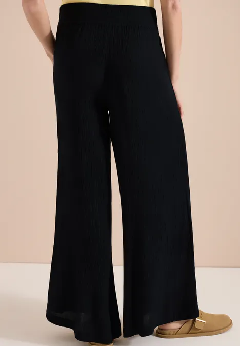 Schwarze, weite Strickhose mit seitlichem Schlitz, dazu goldene, flache Slipper vor neutralem Hintergrund.