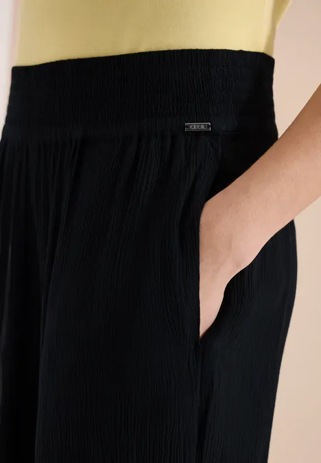 Schwarze, plissierte Hose mit elastischem Bund und seitlicher Eingrifftasche, Hand in Tasche.