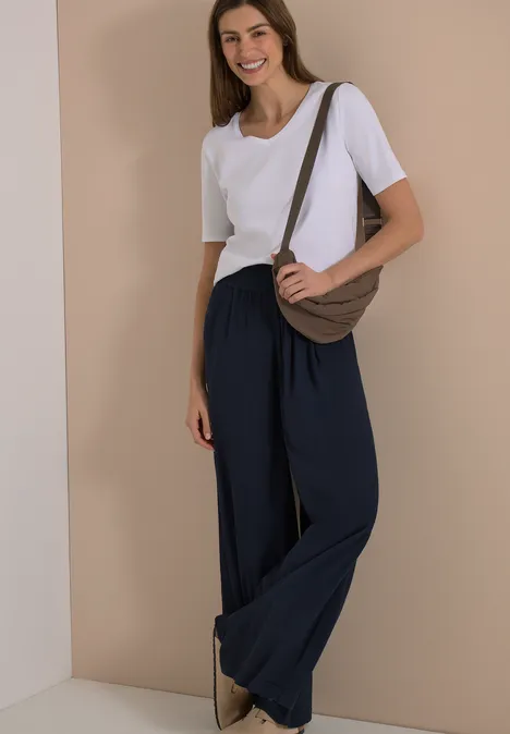 Femme en t-shirt blanc, pantalon large bleu marine et sac à bandoulière, fond sobre.