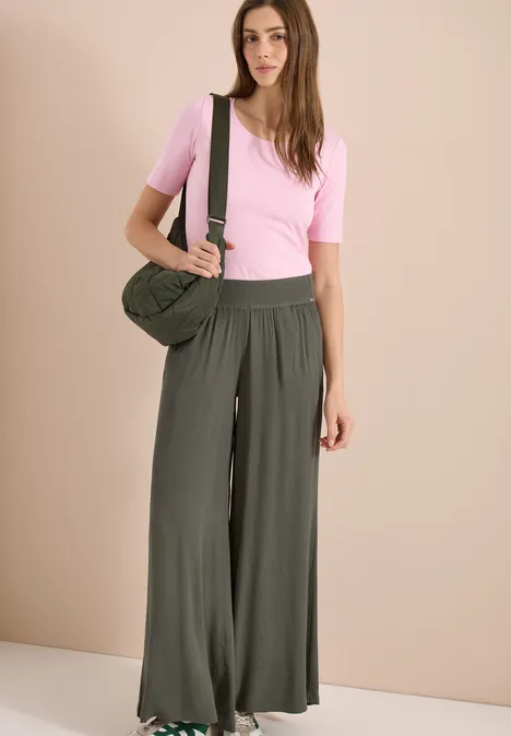 Femme portant un t-shirt rose, un pantalon ample vert et un sac bandoulière sur un fond neutre.