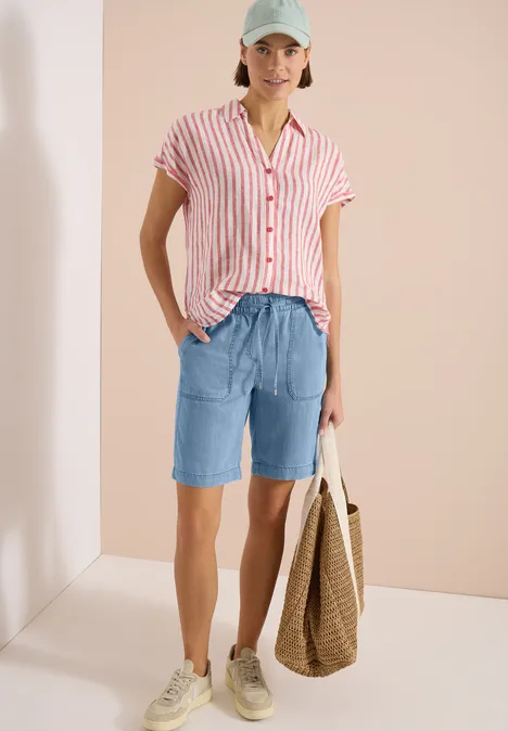 Frau in rosa-weiß gestreiftem Kurzarmhemd, hellblauen Bermudashorts, Stoffbeutel und Sneakers vor neutralem Hintergrund.