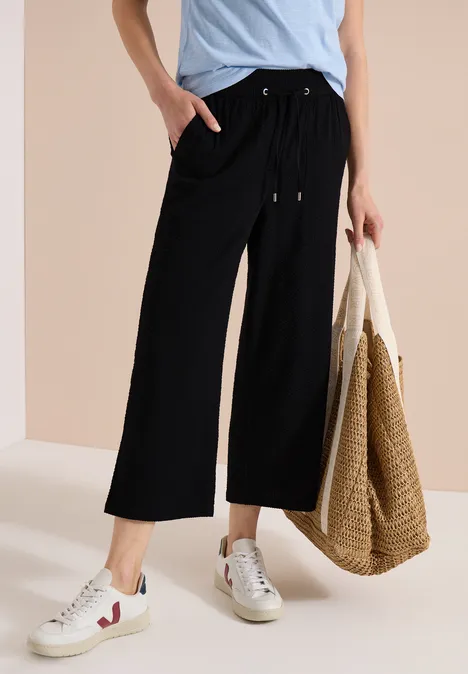 Schwarze Culotte-Hose mit Tunnelzug und weitem Bein, kombiniert mit Sneakern und Strandtasche.